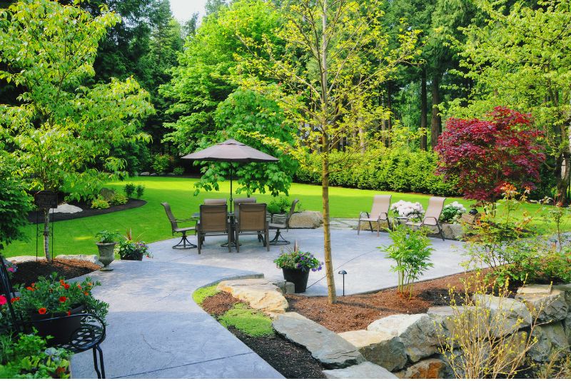 An outdoor picture with a sitting area with chairs and shade. Trees and shrubs are planted